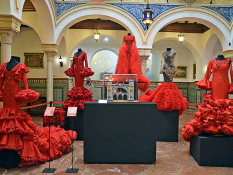 Exposición de vestidos de flamenca rojos en un salón con arcos y lámparas de techo.