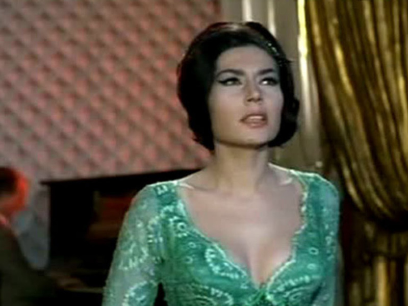 Una mujer vestida con un vestido verde con bordados en la parte superior, posando con una expresión seria en un ambiente de lujo con un hombre en el fondo.