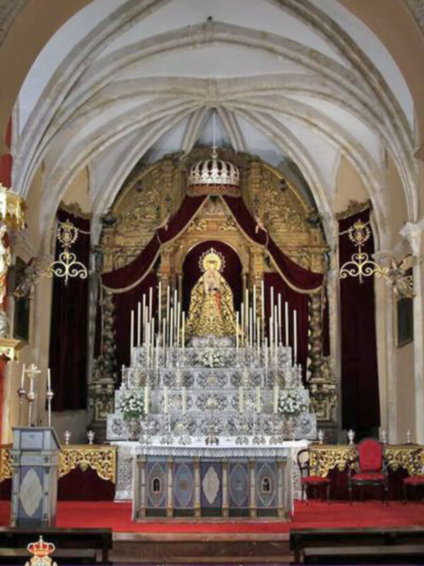Altar con estatua central, adornos dorados y rojos en el fondo. Sobre la imagen, una corona real y un escudo con insignias heráldicas.
