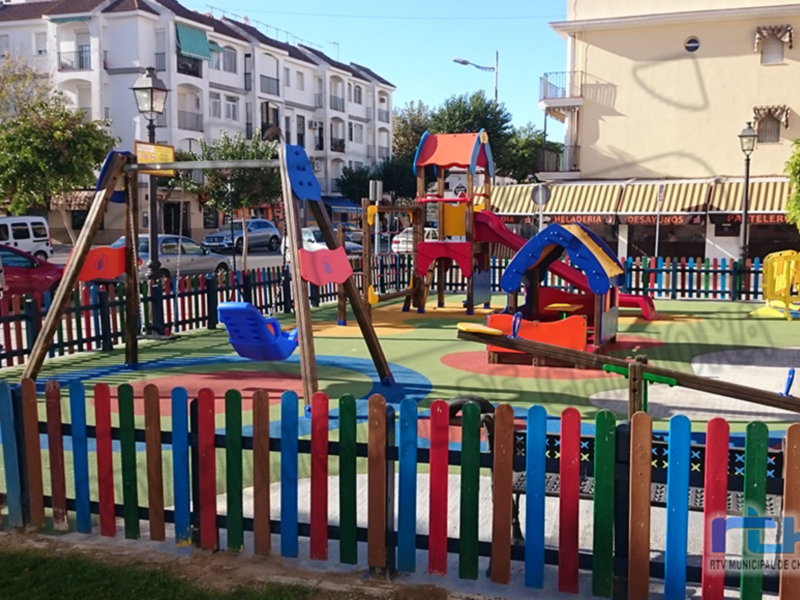 Parque infantil con estructuras de juegos coloridas y un muro de cerca multicolor en el centro, rodeado de árboles y edificios. El cielo es azul claro y hay algunas personas observando el parque.
