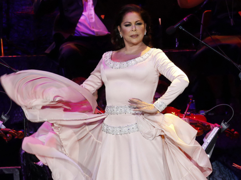 Artista en escenario con vestido blanco elegante, tocando el piano en un concierto con orquesta en el fondo.