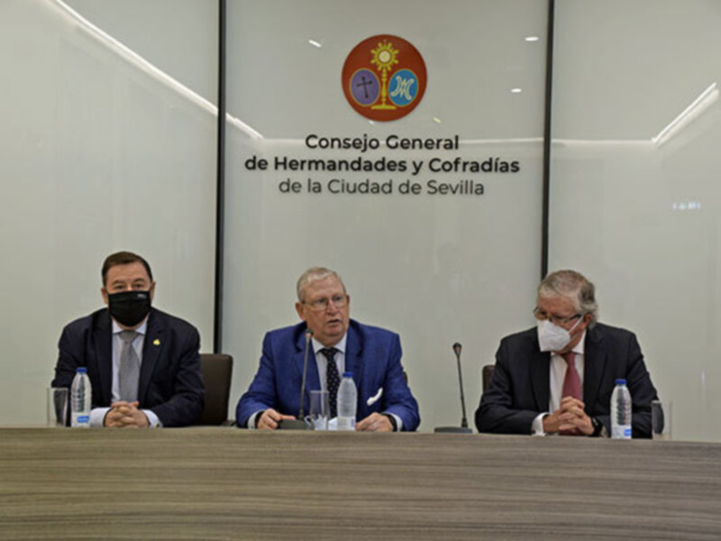 Tres hombres sentados en una mesa con un logo de la Ciudad de Sevilla en el fondo.
