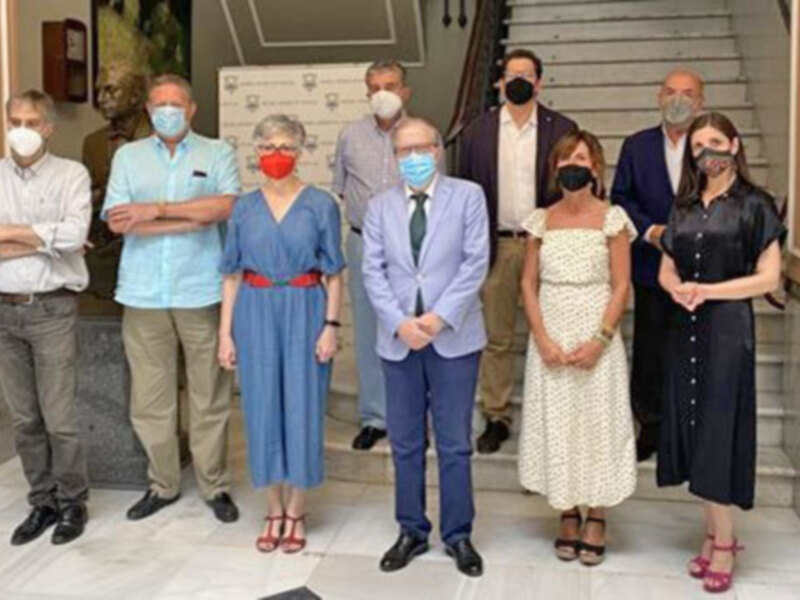 Grupo de personas en una reunión formal, todos con mascarillas, posando en un salón con escalera y decoración clásica.