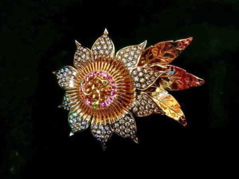 Una broche de diamantes en forma de flor con hojas doradas y detalles en colores.