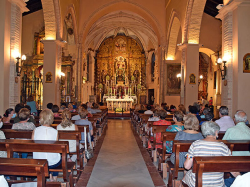 Interior de una iglesia con personas sentadas en bancos, mirando hacia el altar. Decoración barroca y arco de entrada destacados.