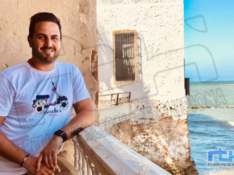Hombre sonriendo en un balcón con vista al mar y edificio antiguo.