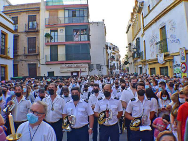 Desfile de músicos con uniformes y máscaras en una calle de Sevilla, con una multitud de espectadores.