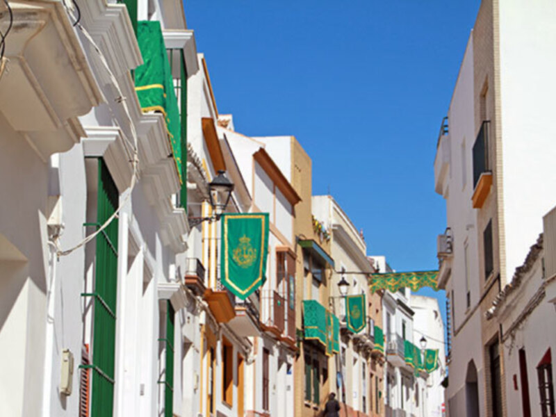 Calle con edificios blancos y balcones, decorados con banderas verdes y amarillas, bajo un cielo azul claro.