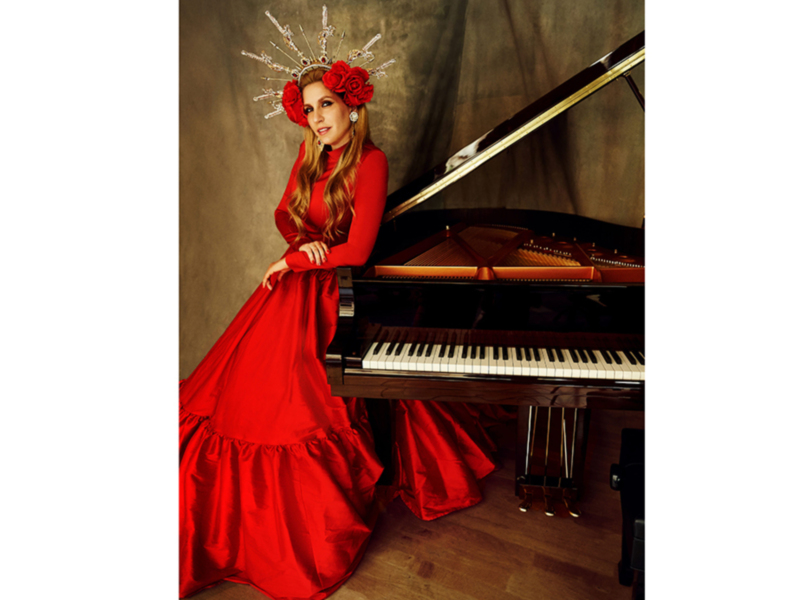 Una mujer vestida de rojo posa junto a un piano, con un tocado de flores en forma de corona en su cabeza.