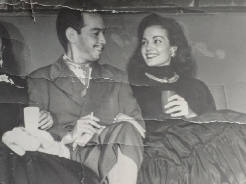 Retrato de pareja en trajes formales, posando juntos con una sonrisa en un entorno interior, vestidos de época.