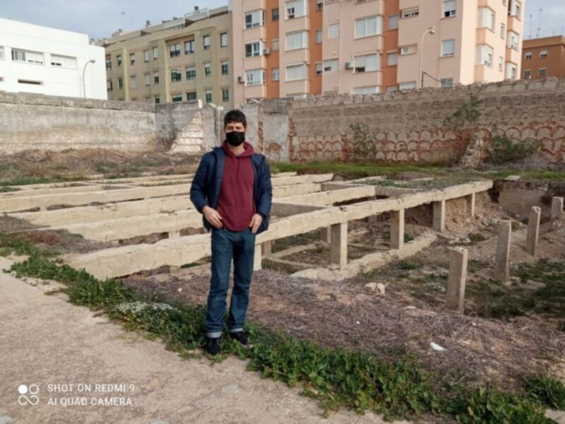 Persona con máscara facial posando frente a estructuras de madera y muros de contención en un entorno urbano.