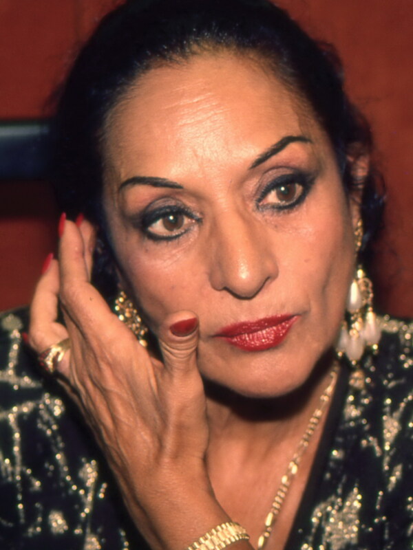 Retrato de mujer con maquillaje dramático y joyas, posando con la mano en el rostro.
