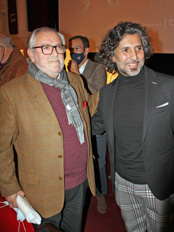 Dos hombres en una recepción formal, uno con chaqueta de tweed y bufanda, el otro con traje oscuro y pantalones a cuadros, posando juntos en un evento cultural.