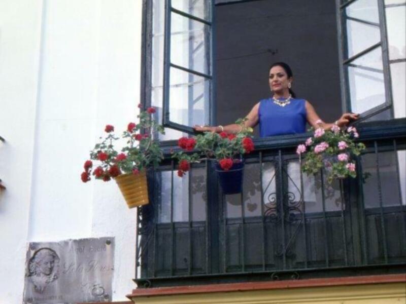 Una mujer en un vestido azul se encuentra en un balcón con flores rojas y un letrero en el fondo. La imagen parece ser de una celebración o evento público.
