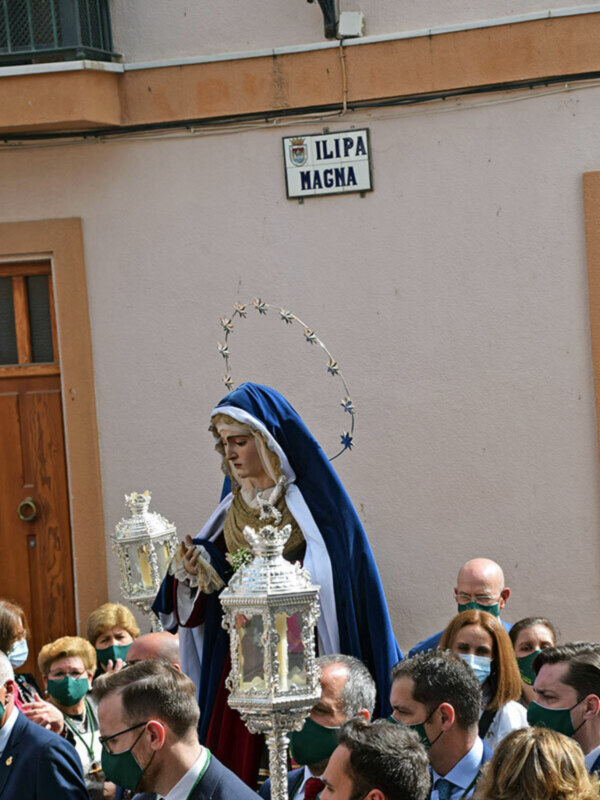 Estatua de la Virgen María en procesión, con una multitud de personas alrededor, en un entorno urbano con una placa que dice 'ILIPA MAGNA' en el fondo.