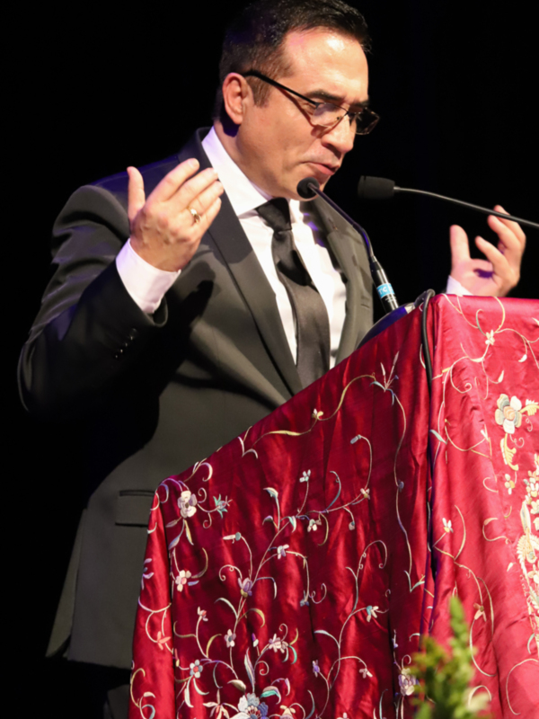 Hombre en traje hablando en un evento formal, detrás de un podio con tela bordada roja.