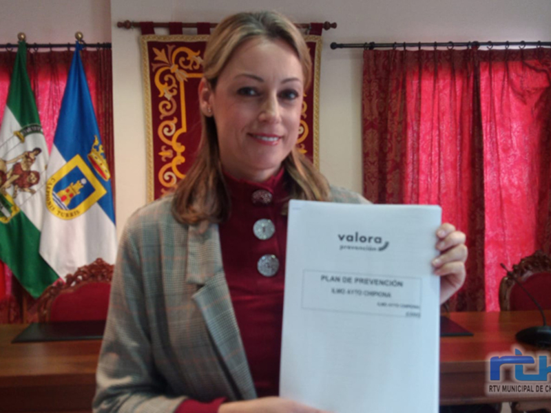 Una mujer sostiene un documento con el título 'Plan de prevención' en un entorno formal, con banderas y cortinas de fondo.