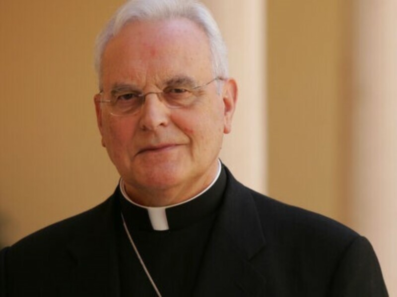Retrato de un sacerdote con vestimenta clerical y gafas, frente a un fondo neutro.