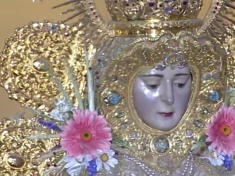 Imagen de una imagen de la Virgen María adornada con joyas y flores, destacando su majestuosidad y belleza.