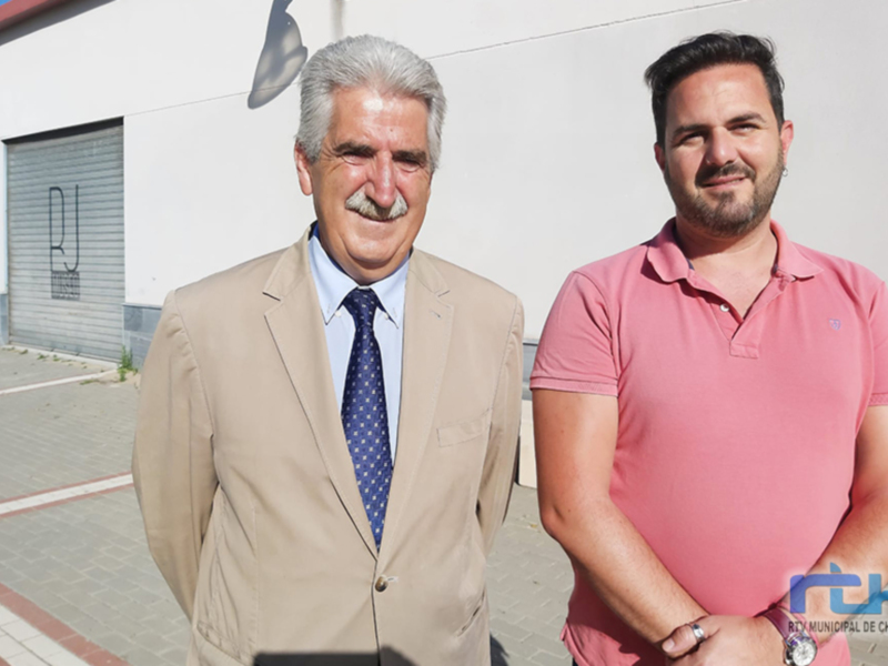 Dos hombres posan juntos al aire libre, uno en traje y otro en camiseta rosa, frente a un edificio industrial.