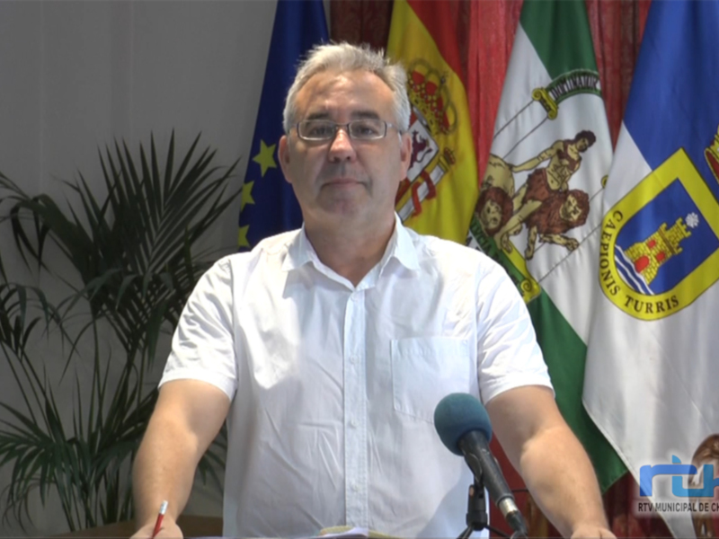 Un hombre habla en una conferencia de prensa en Chipiona, con banderas españolas y de Chipiona en el fondo.