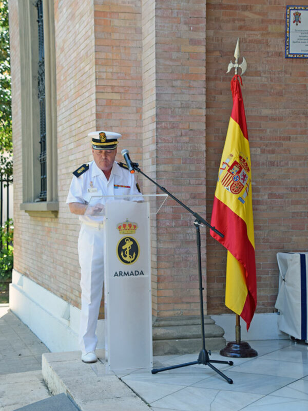 Un oficial de la Armada Española pronuncia un discurso en un evento oficial, con la bandera española y el escudo naval en el fondo.
