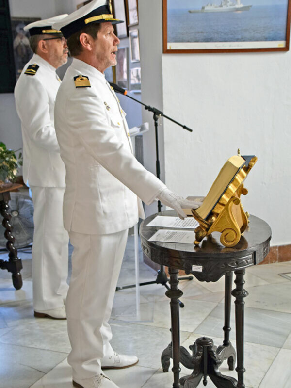 Dos oficiales militares en uniforme blanco, uno de ellos sostiene un libro de texto, en un entorno formal con un paisaje marítimo en el fondo.