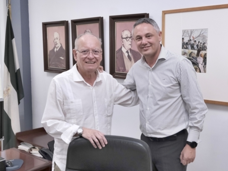 Dos hombres sonrientes en un entorno profesional, rodeados de retratos y banderas.