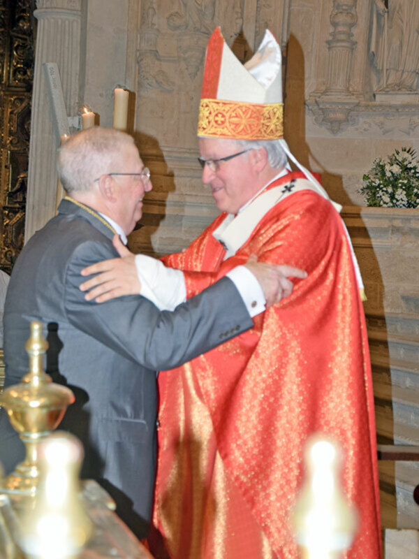 Un obispo en vestimenta sacerdotal roja abraza a un hombre en traje formal en un ambiente de iglesia adornada.