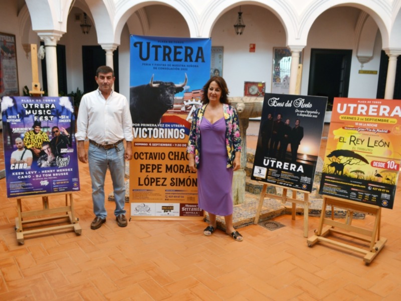 Un hombre y una mujer posan junto a varios carteles promocionando eventos culturales en Utrera, Sevilla. Los carteles incluyen información sobre festivales de música, conciertos y otros eventos locales.
