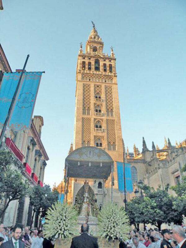 La Giralda, emblemático campanario de la Catedral de Sevilla, bajo un cielo claro y con una procesión en el primer plano.