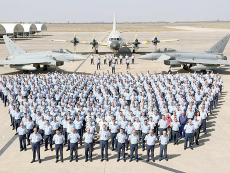 Grupo de personal uniformado en una base aérea con aviones de combate en el fondo.