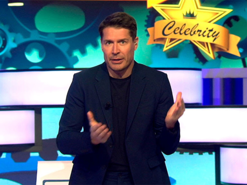 Presentador en un escenario con fondo de engranajes y la palabra "Celebrity" en un estandarte dorado.