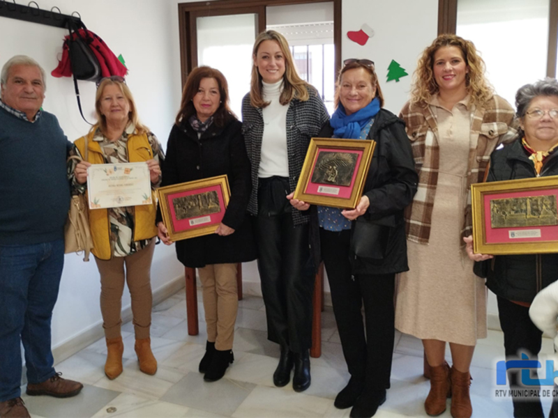 Una imagen de un grupo de personas posando con sus premios, incluyendo una mujer en el centro que sostiene un certificado y dos cuadros dorados. La escena parece ser en una oficina o sala de conferencias, con decoraciones navideñas al fondo.
