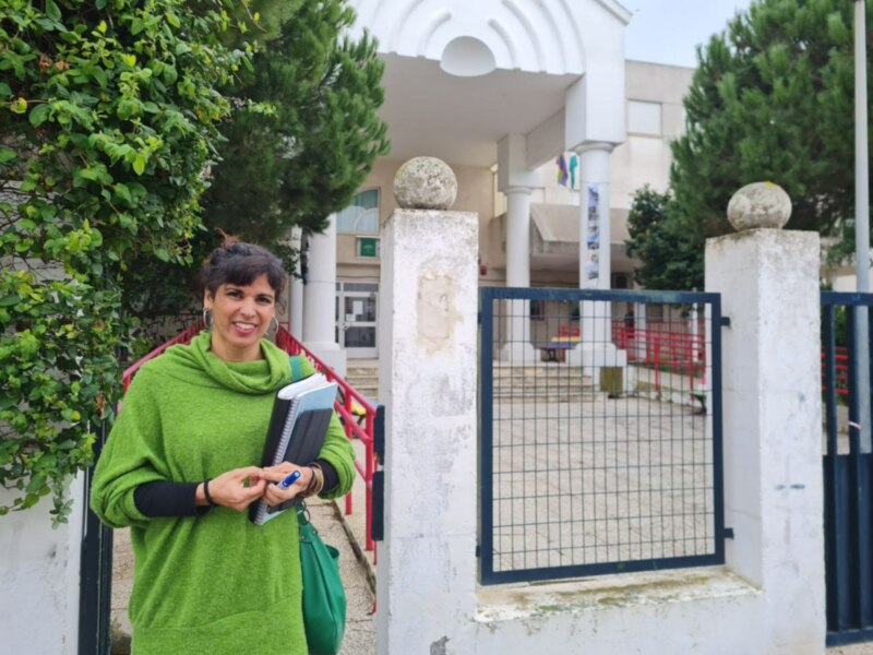 Mujer sonriente con cuaderno y bolígrafo en la entrada de un edificio educativo