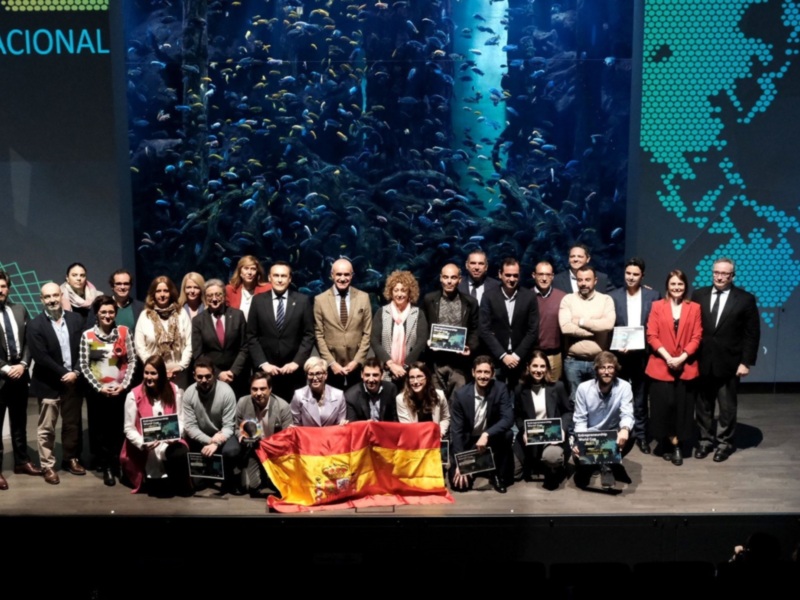 Grupo de personas en una ceremonia de premiación en España, con la bandera española y un fondo de acuario.