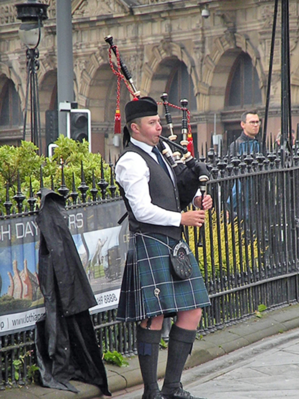Un músico escocés toca el uilleann pipes en una parada frente a un edificio histórico con arcos. Detalles como el vestido tradicional escocés, la chaqueta de cuero y los guantes negros son visibles.