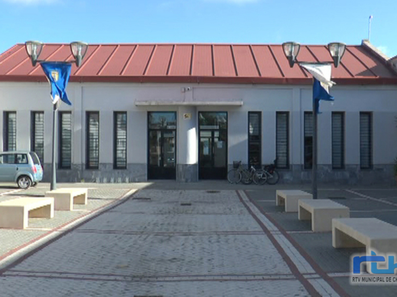Fachada de un edificio municipal con una bandera azul y blanca en el exterior. El edificio tiene una cubierta roja y está rodeado de bancos de hormigón y bicicletas aparcadas en el exterior.