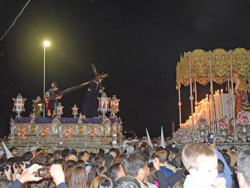 Procesión nocturna con santos en procesión, iluminada por candelabros y espectadores capturando el momento.