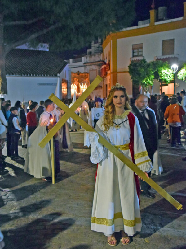 Procesión nocturna con vestimenta tradicional y cruz, en un entorno urbano iluminado.