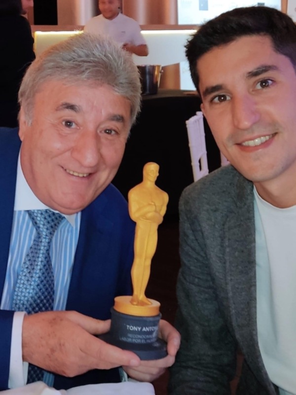 Dos hombres sonrientes posan con un trofeo de reconocimiento en un evento formal.