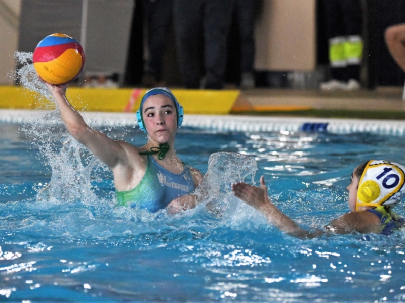Jugadores de waterpolo en acción en una piscina, con una pelota en el aire y una figura en el fondo.