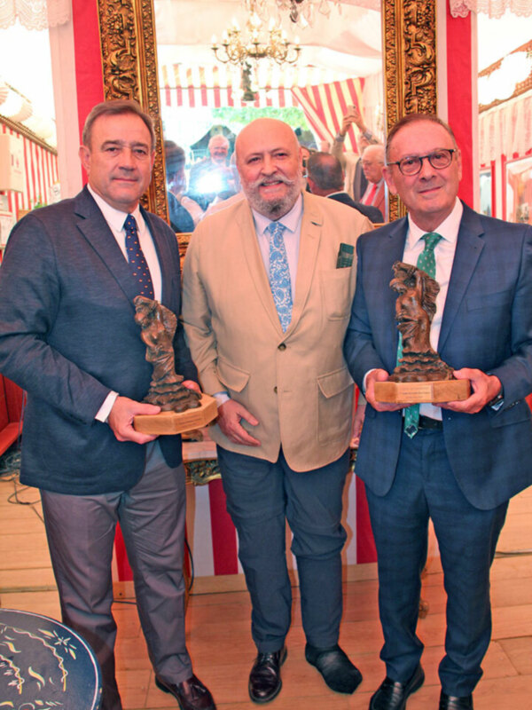 Tres hombres en trajes celebran con trofeos en una recepción formal.