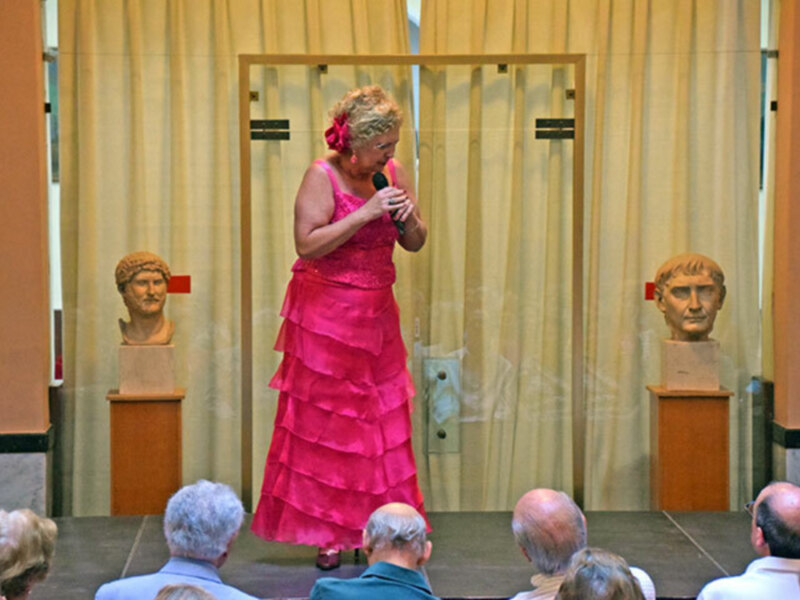 Mujer en vestido rosa hablando en un escenario con bustos antiguos al fondo.