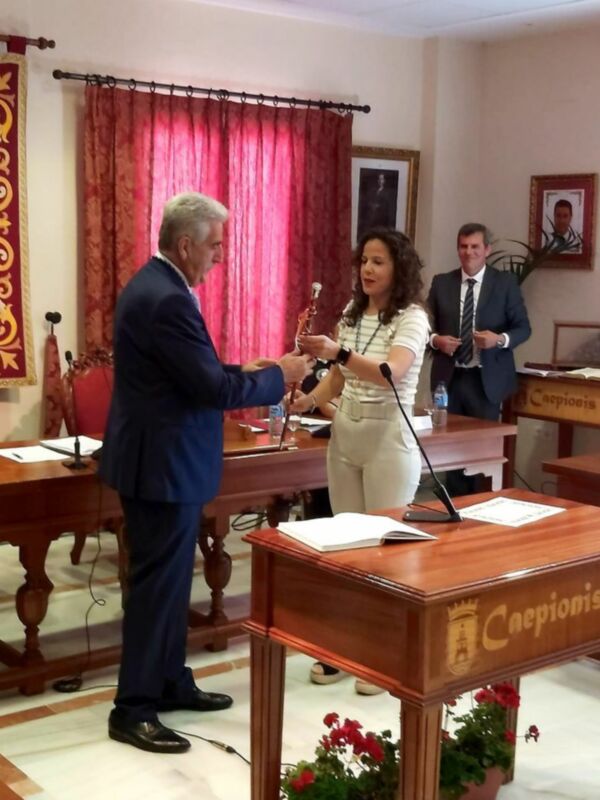 En un ambiente formal, una mujer recibe un reconocimiento de manos de un hombre en traje. La escena se desarrolla en una sala con cortinas rojas y un escritorio de madera. Detalles como el logo "Caepionis" en la mesa y las flores rojas en el suelo añaden contexto al ambiente.