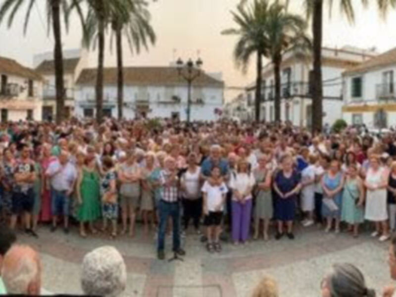 Gente reunida en una plaza con palmeras y edificios históricos al fondo.
