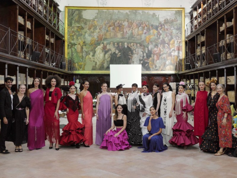 Desfile de moda flamenca en un espacio biblioteca con un mural histórico en el fondo.