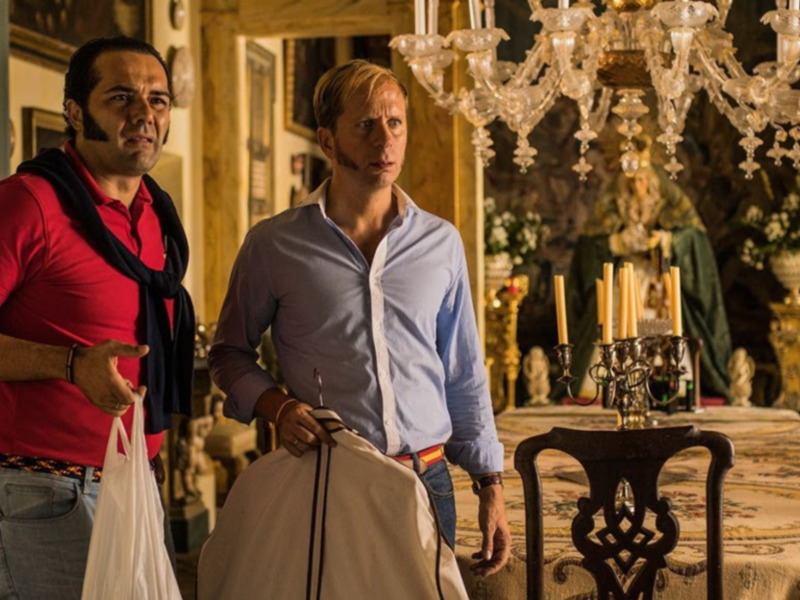 Dos hombres en una sala de estilo antiguo, uno con una bolsa de plástico, parecen estar en una conversación seria.