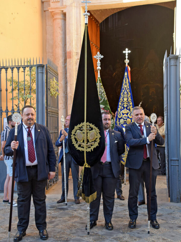 Oficiales de una procesión religiosa frente a una iglesia, sosteniendo estandartes y palmas.