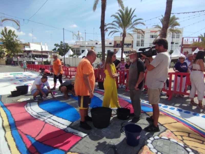 Un grupo de personas pintando un tapete de colores en una calle, con palmeras y edificios al fondo. Un hombre con una cámara filmando el evento.